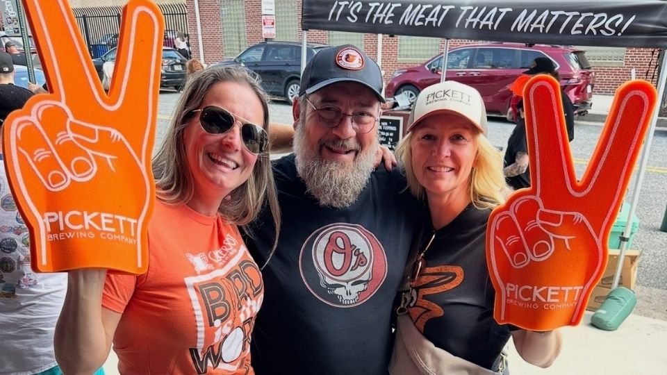 Three people smiling and holding orange Pickett Brewing Company foam fingers at an outdoor event.